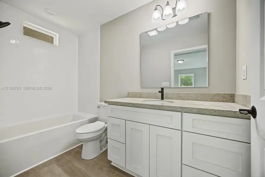 Bathroom featuring a vanity with white shaker-style cabinetry, neutral stone-finish countertop, undermount sink with dark metal hardware, and wood-finish flooring, complemented by a tiled shower surround Bathroom featuring a vanity with white shaker-style cabinetry, neutral stone-finish countertop, undermount sink with dark metal hardware, and wood-finish flooring, complemented by a tiled shower surround