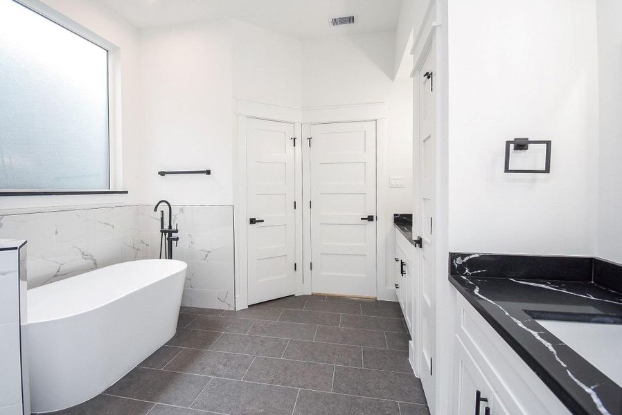 This bathroom features a modern design with a freestanding tub, sleek black fixtures, and a dual vanity with black countertops. It has large tile flooring and a frosted window for natural light and privacy.