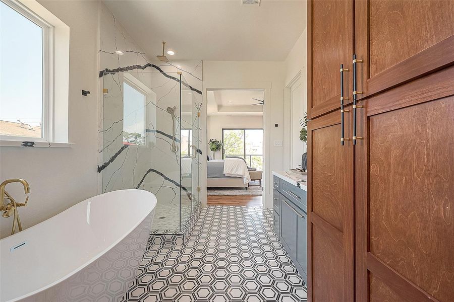 This is a modern bathroom featuring a standalone bathtub, a glass-enclosed shower with marble walls, and an eye-catching geometric floor tile design. There is a view into the adjoining bedroom, indicating an en-suite layout. This is a modern bathroom featuring a standalone bathtub, a glass-enclosed shower with marble walls, and an eye-catching geometric floor tile design. There is a view into the adjoining bedroom, indicating an en-suite layout.