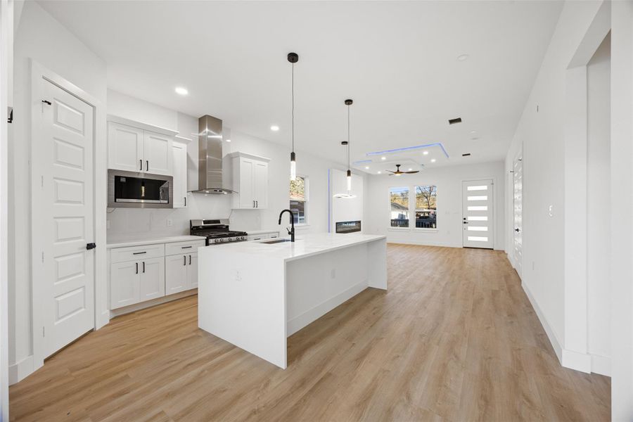 This modern open-concept kitchen and living area features sleek white cabinetry, a large island with pendant lighting, stainless steel appliances, and wood flooring. The space is bright and airy, with a contemporary ceiling fan and ample natural light from large windows.