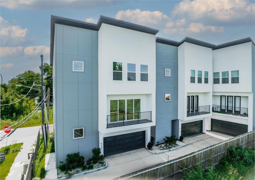 Modern townhouses with a sleek design, featuring large windows and balconies. Each unit includes a spacious garage. The exterior combines white and gray panels, set against a backdrop of greenery and a clear sky.