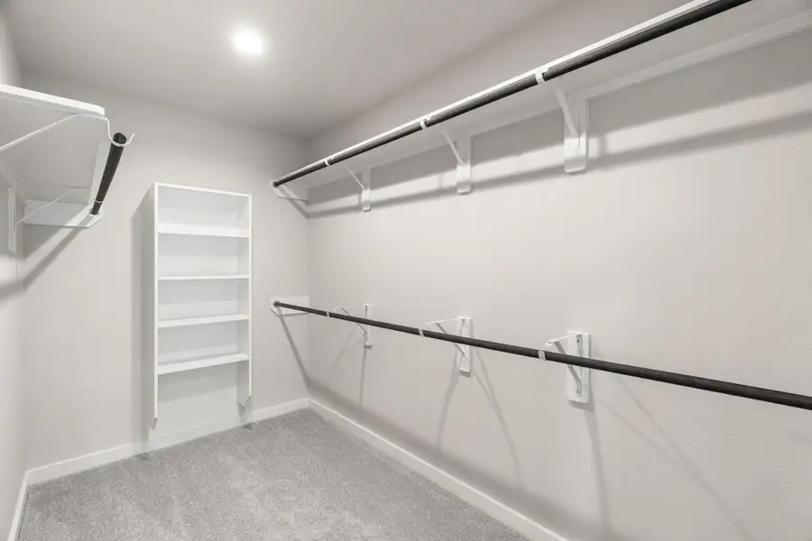 Experience luxury in this spacious walk-in closet with high ceilings and plush carpet. Warm paint tones, built-in shelving, and dark finishes create a contemporary and functional retreat. Sample photo of completed home with similar floor plan. As-built interior colors and selections may vary.