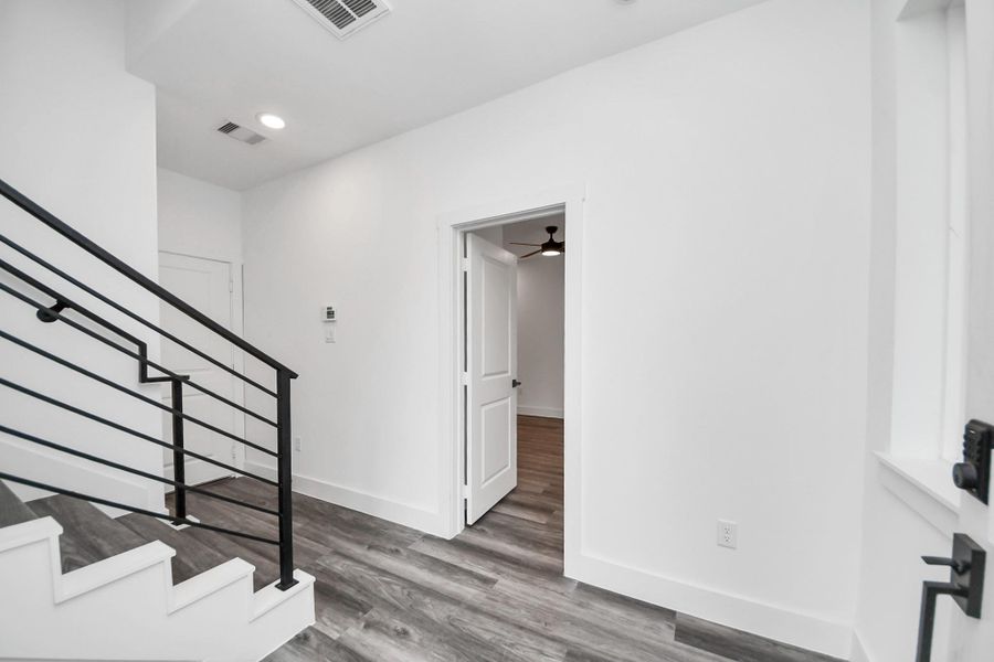 This photo shows a modern entryway with white walls and wood flooring. It features a staircase with sleek black railings, recessed lighting, and a door leading to another room. This photo shows a modern entryway with white walls and wood flooring. It features a staircase with sleek black railings, recessed lighting, and a door leading to another room.