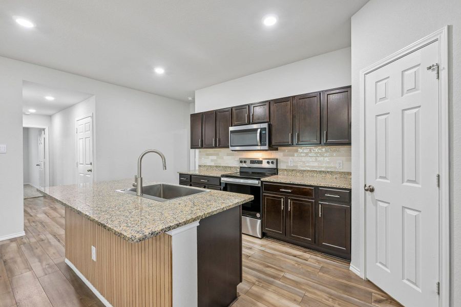 Kitchen featuring a spacious island with a single-basin undermount sink, granite countertops, wood-finish flooring, dark wood cabinetry, and stainless steel appliances including a microwave and range