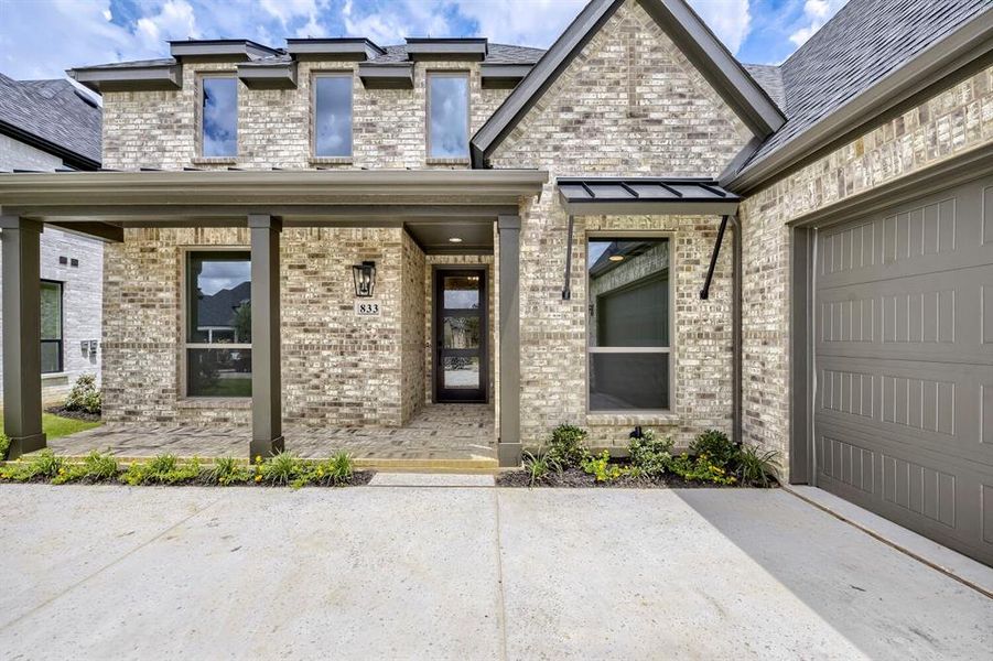 Brick facade featuring a covered entry porch, a modern glass-paneled front door, and a two-car garage with a panel-style door Brick facade featuring a covered entry porch, a modern glass-paneled front door, and a two-car garage with a panel-style door