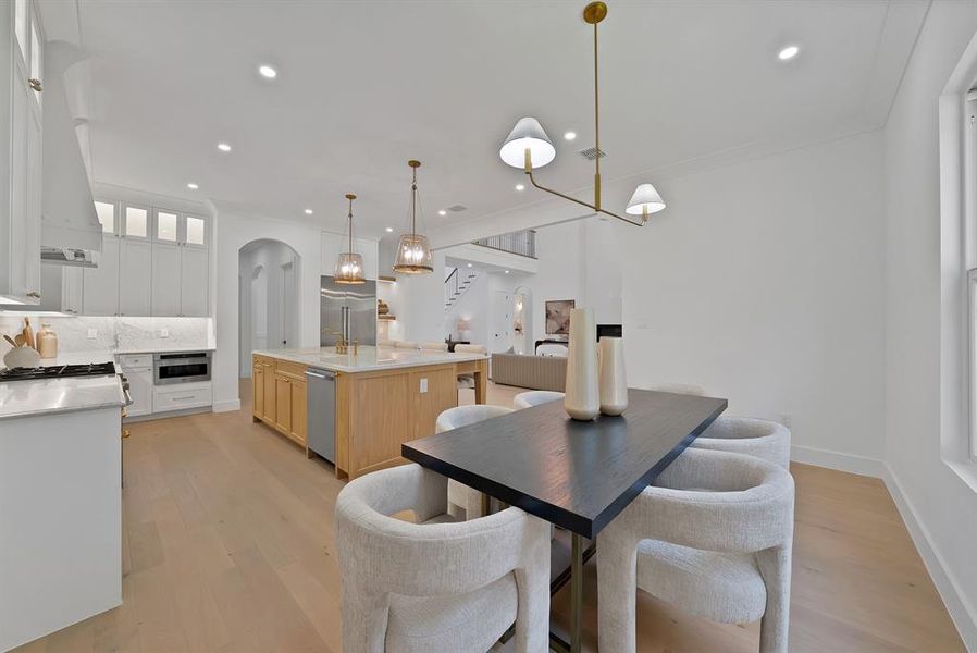 Bright kitchen and dining area featuring light wood-finish flooring, white cabinetry, a large kitchen island with pendant lighting, integrated stainless steel appliances, and a linear chandelier over the dining table