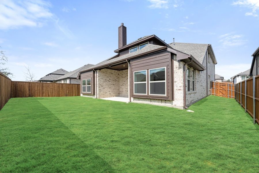 Rear view of brick home with covered patio, large grassy backyard, and wood privacy fence