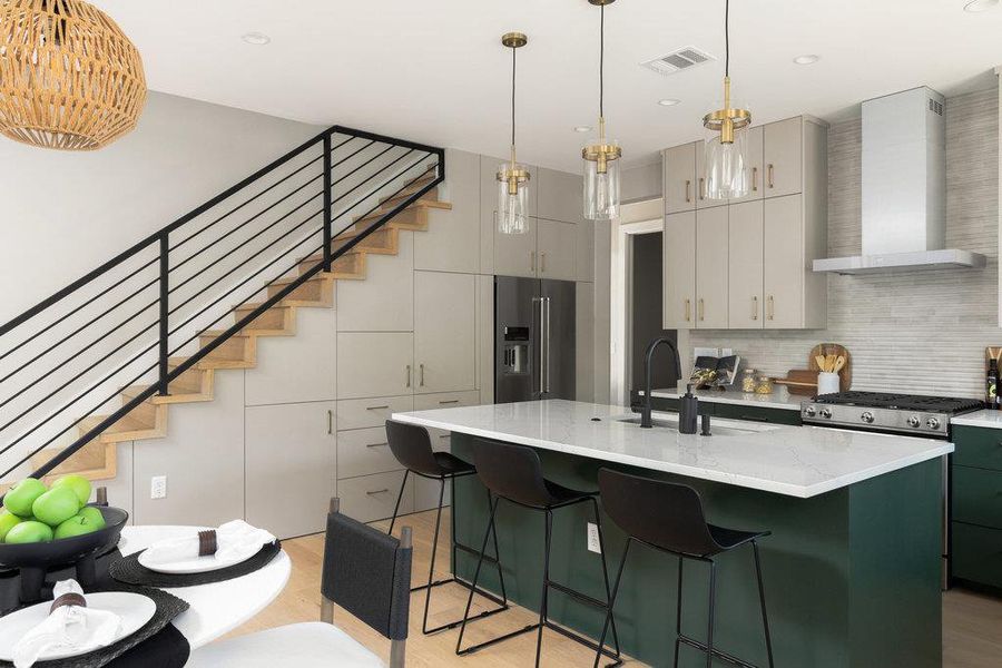 Kitchen with green cabinetry, a center island with sink, high end fridge, wall chimney exhaust hood, and hanging light fixtures