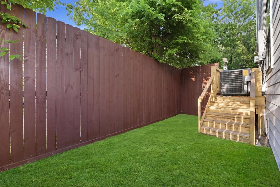 Private, low-maintenance side yard featuring lush turf grass, tall privacy fencing, and wood steps leading to an elevated deck—ideal for outdoor relaxation, pets, or entertaining.