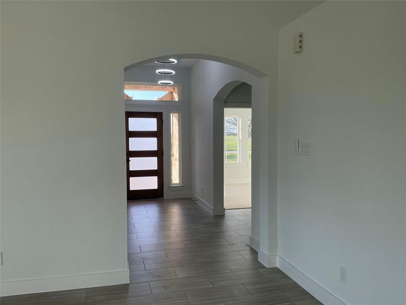 Bright entryway featuring modern tile flooring and a stylish wooden front door with frosted glass panels. Arched openings lead to a well-lit space with large windows showcasing outdoor views.