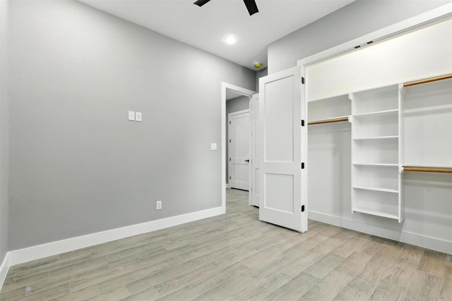 This room features a sleek, modern design with light flooring and neutral gray walls. It includes a spacious walk-in closet with ample shelving and hanging space, and a door leading to an adjacent area. Recessed lighting and a ceiling fan enhance the room's functionality and comfort.