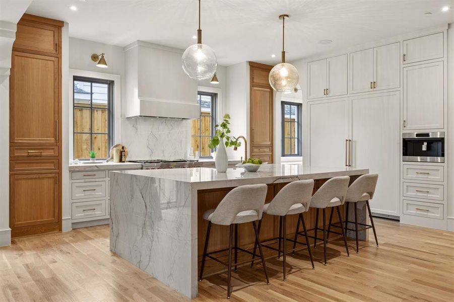 Gourmet kitchen featuring a waterfall island with stone countertop, integrated appliances, natural wood-finish flooring, custom cabinetry, and a stone backsplash Gourmet kitchen featuring a waterfall island with stone countertop, integrated appliances, natural wood-finish flooring, custom cabinetry, and a stone backsplash