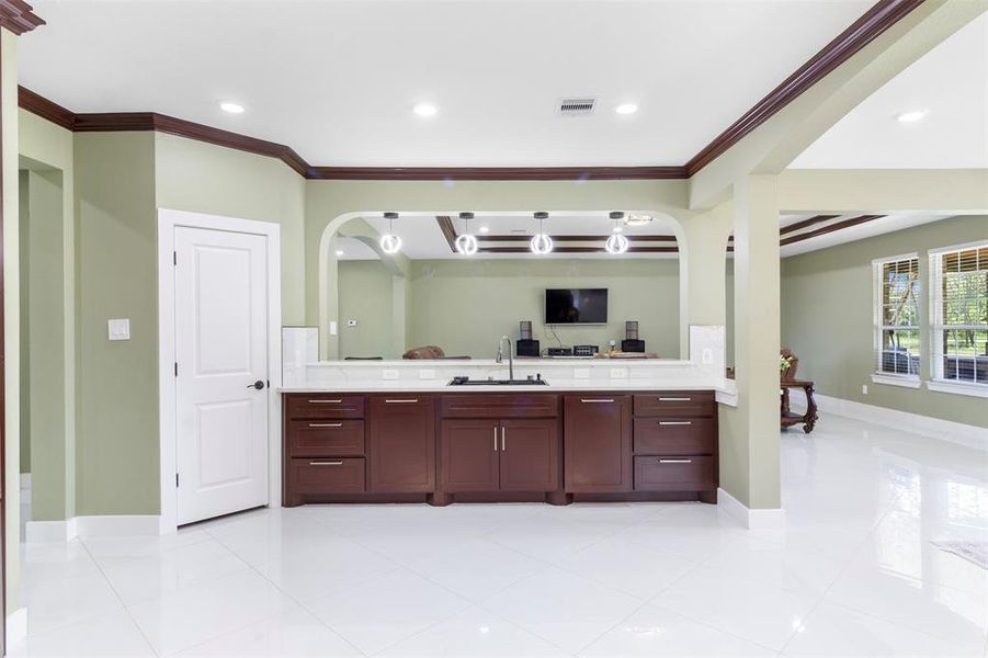 This photo features a spacious open-concept living area with elegant crown molding and recessed lighting. The room has light green walls and a glossy white tile floor. A modern kitchen island with dark wood cabinetry is centrally located, and large windows provide ample natural light.