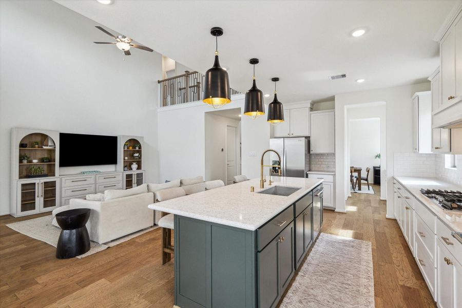 A long view of the kitchen showcases its seamless flow into the living spaces, with the island’s deep green cabinetry, granite countertop, brass hardware, and a convenient coffee nook beside the refrigerator/freezer.