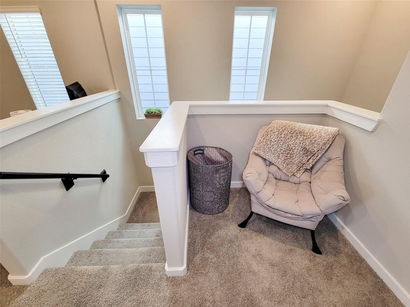 This cozy landing area features carpeted stairs, a comfortable chair with a throw, and natural light from tall windows, creating a welcoming nook.