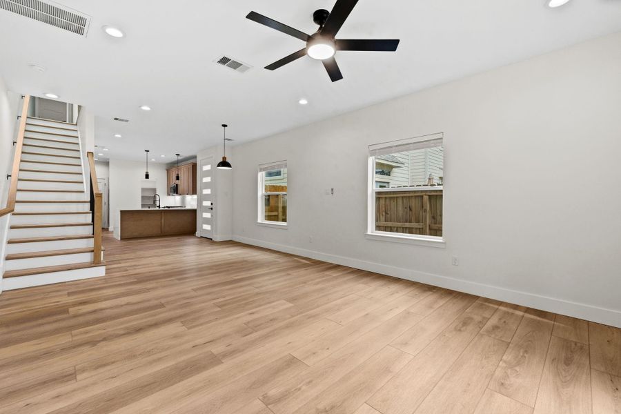 Spacious open living area with warm wood flooring, modern ceiling fan, and abundant natural light, seamlessly connected to a sleek kitchen and dining space.