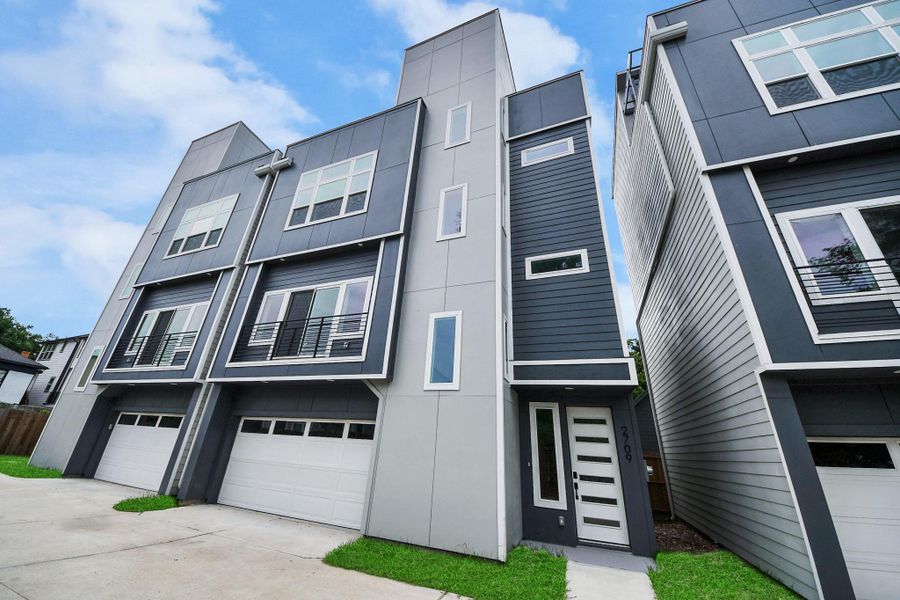 This is a modern, four-story townhouse with a sleek, geometric design. It features large windows, a two-car garage, and a contemporary facade with contrasting dark and light panels. The property is situated in a residential area with a small lawn at the front. This is a modern, four-story townhouse with a sleek, geometric design. It features large windows, a two-car garage, and a contemporary facade with contrasting dark and light panels. The property is situated in a residential area with a small lawn at the front.