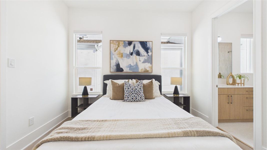 This bright bedroom features a cozy bed with elegant pillows and a modern abstract painting. It has two bedside tables with lamps and large windows for natural light. An ensuite bathroom is visible, offering convenience and style with its sleek vanity and decor.