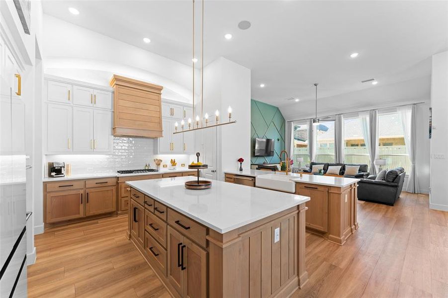 The kitchen features a large island with a white countertop and natural wood cabinetry, a white subway tile backsplash, and a gold-toned chandelier
