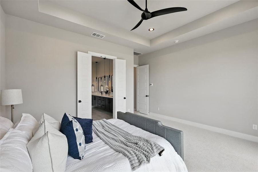 Carpeted bedroom with a raised ceiling, recessed lighting, connected bathroom, and ceiling fan