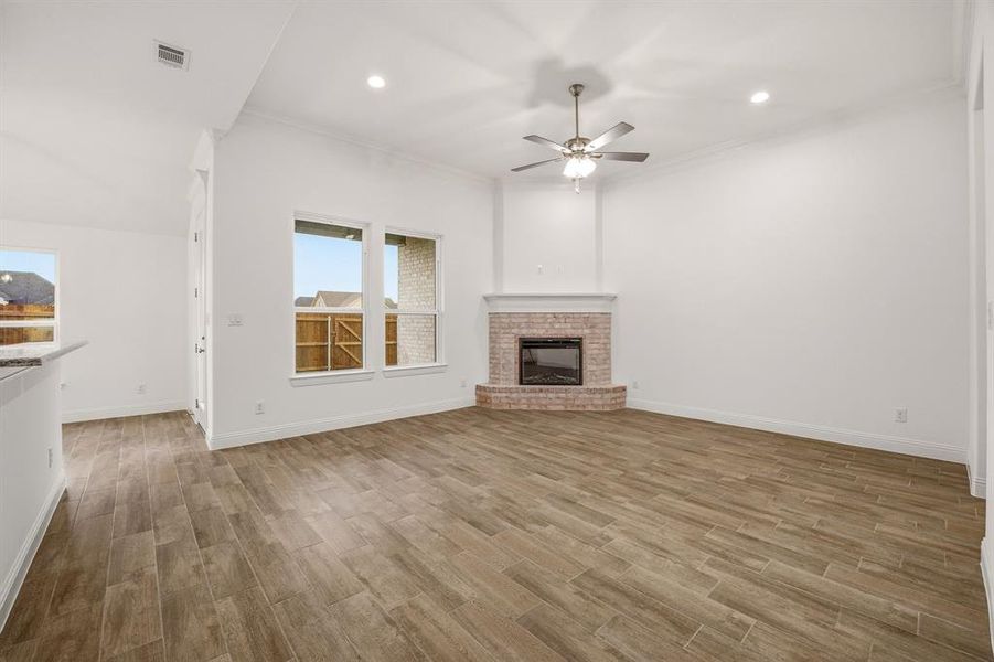 Spacious living area featuring light wood-look flooring, a corner brick fireplace, and a ceiling fan with integrated lighting