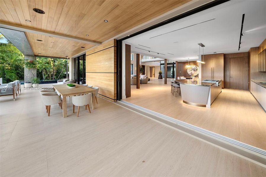Seamless indoor-outdoor connection via pocket sliding glass doors opening from chef's kitchen and dining area to covered wood-ceiling loggia with outdoor teak dining table, woven chairs, and pool terrace beyond.
