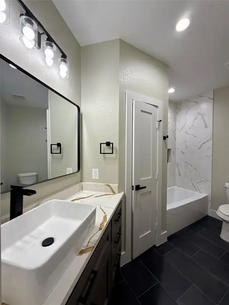 This modern bathroom features a sleek design with a large rectangular sink, Quartz countertop, and elegant black fixtures. The room includes a bathtub with a marble-style wall and dark tile flooring, creating a sophisticated contrast.