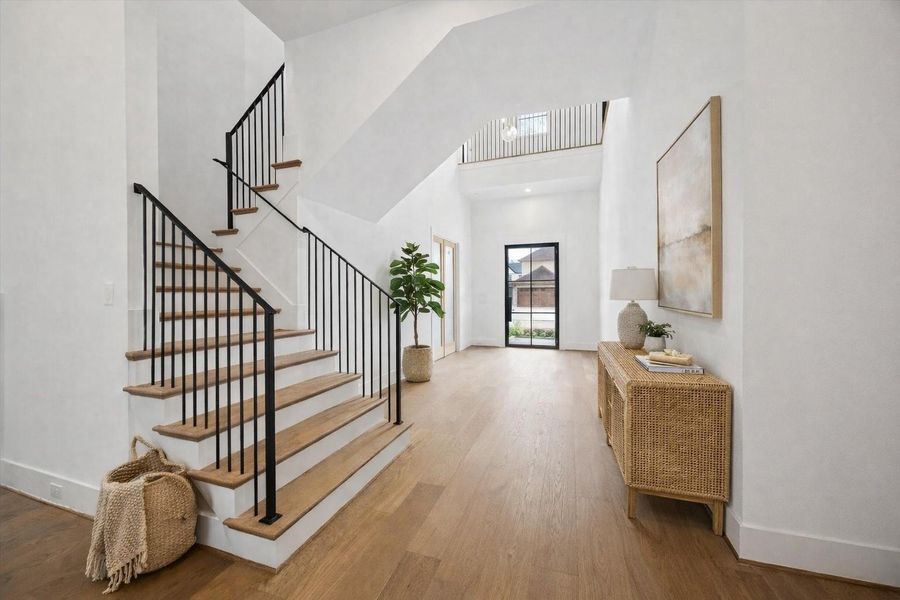 Soaring ceilings and a modern floating staircase create a dramatic first impression in this light filled entry. Warm oak treads, black iron balusters, and wide plank hardwood floors pair with curated staging to highlight the open concept layout and seamless flow to the front glass door, offering both architectural impact and everyday livability. *Virtually Staged*