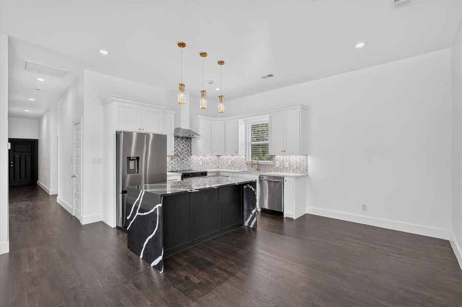 Open-concept kitchen featuring an oversized island with waterfall-edge countertop, stainless steel appliances, white shaker cabinetry, and a chevron tile backsplash