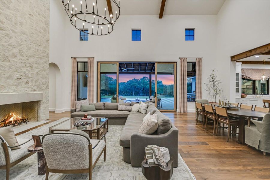 Living area featuring a chandelier, beam ceiling, light wood-style flooring, a towering ceiling, and a large fireplace Living area featuring a chandelier, beam ceiling, light wood-style flooring, a towering ceiling, and a large fireplace