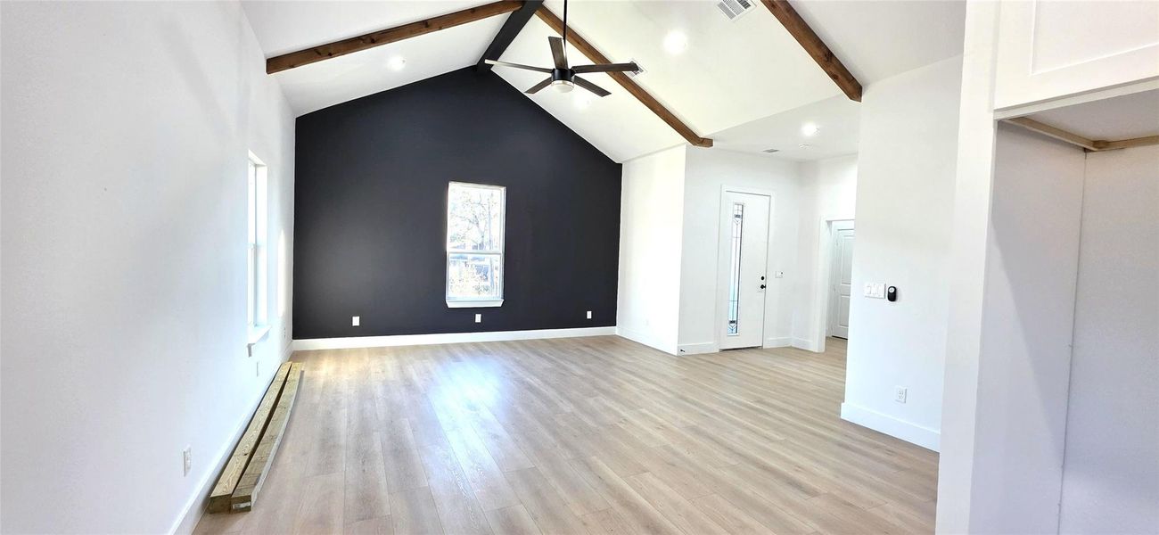 This spacious room features a vaulted ceiling with beams, a sleek ceiling fan, and a striking dark accent wall. Natural light streams in through a central window, highlighting the light flooring. The space feels modern and airy, perfect for versatile living arrangements.