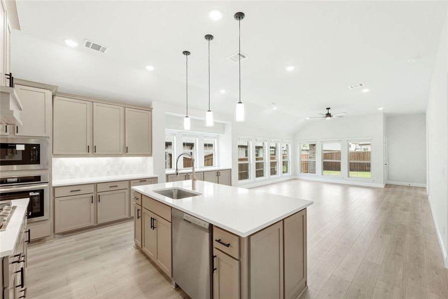 Open kitchen with gray cabinets, center island with sink, pendant lights, and light wood floors opening to living area
