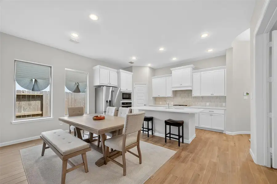 This photo showcases a bright, open-concept kitchen and dining area. It features white cabinetry, modern appliances, and a large island with seating. The space is well-lit with recessed lighting and natural light from several windows, complemented by light wood look flooring. This photo showcases a bright, open-concept kitchen and dining area. It features white cabinetry, modern appliances, and a large island with seating. The space is well-lit with recessed lighting and natural light from several windows, complemented by light wood look flooring.