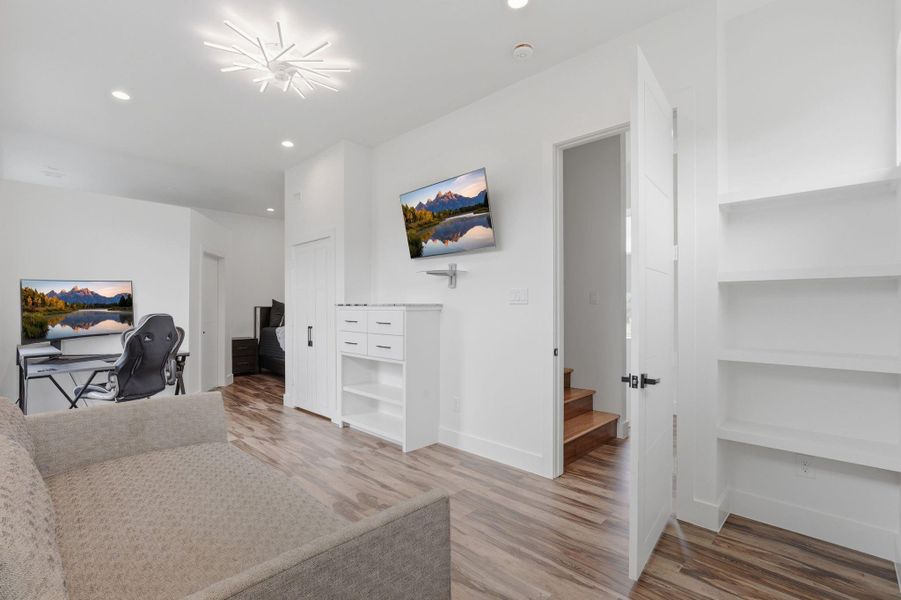 This room features a modern design with sleek hardwood floors, built-in shelving, and a contemporary light fixture. It includes a cozy seating area, a workspace with a desk and gaming chair, and wall-mounted TVs. The open door leads to a staircase with access to additional spaces.