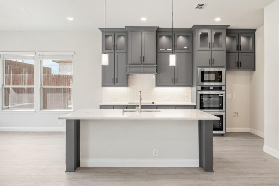 Kitchen with gray cabinets, white island with sink, double wall oven, and pendant lights over light wood floors