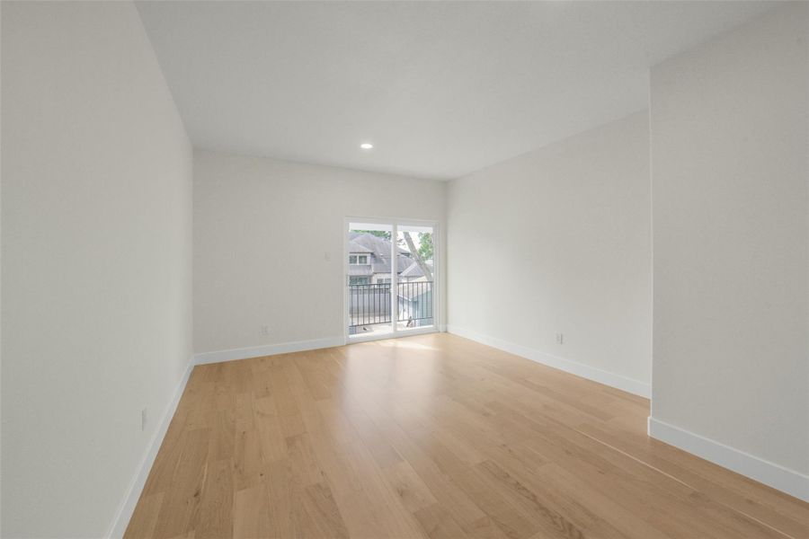 Bright and spacious room with light wood flooring, neutral walls, and a sliding glass door leading to a small balcony with a neighborhood view.