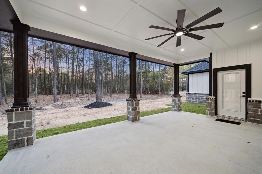 View from the rear of the home toward the lot.  Trees with mulch surround.   Covered rear patio shows the stone and wood support beams of the patio.  Door with full light glass opens to the dining room.  Door also has glass enclosed blinds.   Recessed lighting and large lighted ceiling fan.