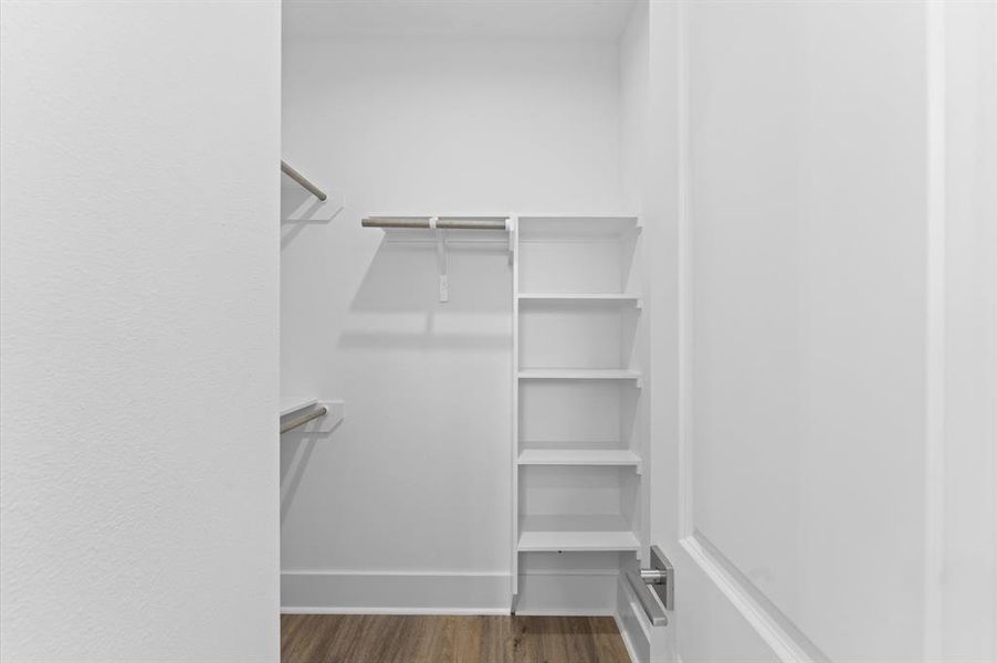 This photo shows a bright, walk-in closet with white walls and shelving. It features multiple hanging rods and built-in shelves, offering ample storage space. The flooring is a warm wood tone, creating a clean and organized feel.