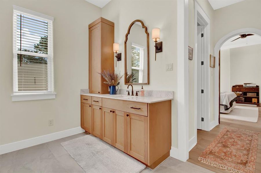 The primary bathroom features identical wooden vanities with marble countertops, a large mirror, and elegant wall sconces. A window provides natural light, and there's an arched doorway leading to the adjoining bedroom.