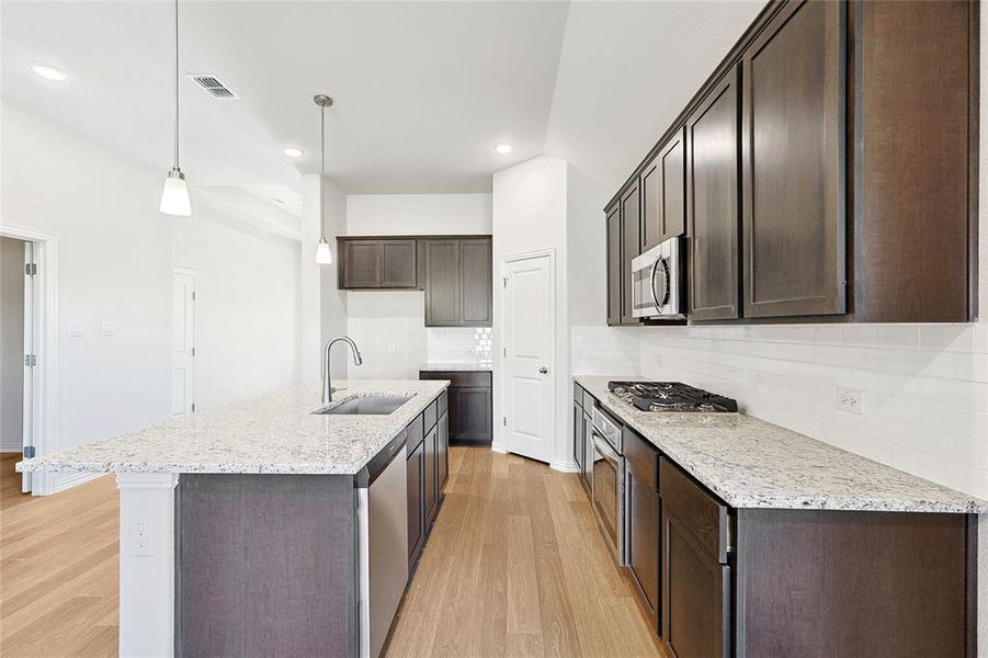 Kitchen featuring decorative light fixtures, tasteful backsplash, stainless steel appliances, light hardwood / wood-style floors, and a kitchen island with sink