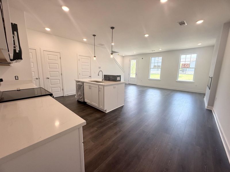 Open-concept living space featuring wood-finish flooring, recessed lighting, a modern kitchen island with sink, white shaker cabinetry, and light-toned countertops