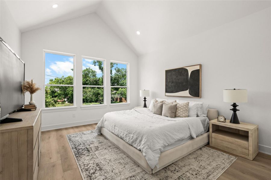 The primary bedroom features a dramatic vaulted ceiling and three large vertical windows that look out onto mature trees. The spacious layout easily fits a king-sized bed, matching nightstands, and a large dresser with room to spare.