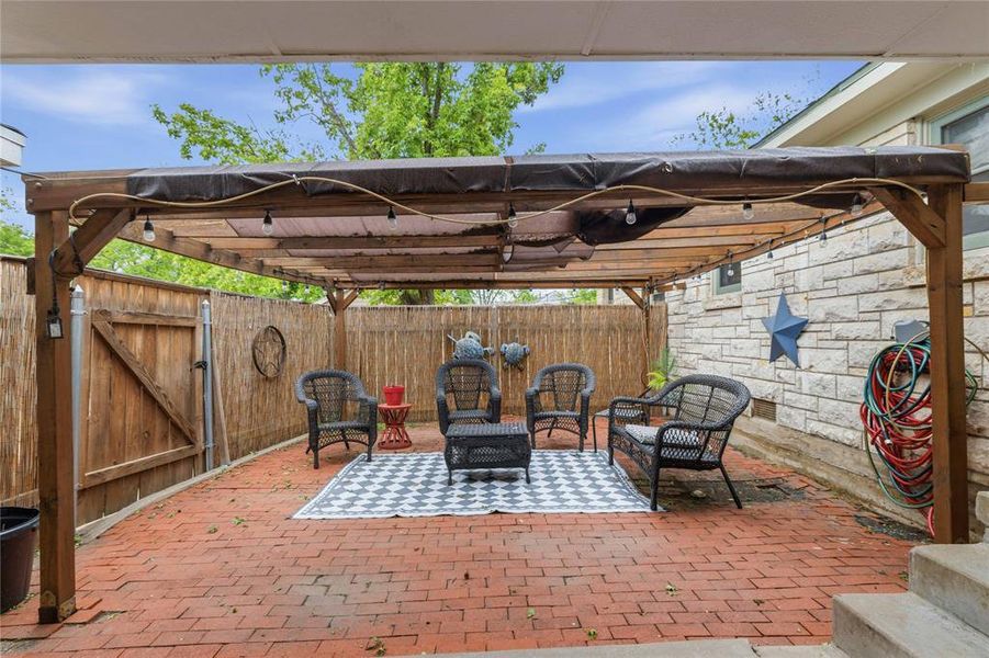 Outdoor patio area featuring a wooden pergola with string lighting, a red brick-finish patio floor, stone-veneer exterior wall, and a wood privacy fence Outdoor patio area featuring a wooden pergola with string lighting, a red brick-finish patio floor, stone-veneer exterior wall, and a wood privacy fence