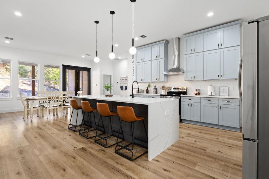 Kitchen featuring stainless steel appliances, a kitchen island with sink, pendant lighting, light stone countertops, and wall chimney exhaust hood