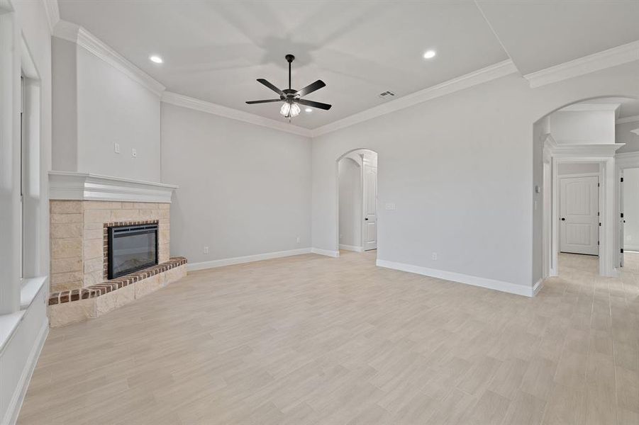 Unfurnished living room featuring light wood-style flooring, arched walkways, crown molding, a brick fireplace, and a ceiling fan Unfurnished living room featuring light wood-style flooring, arched walkways, crown molding, a brick fireplace, and a ceiling fan