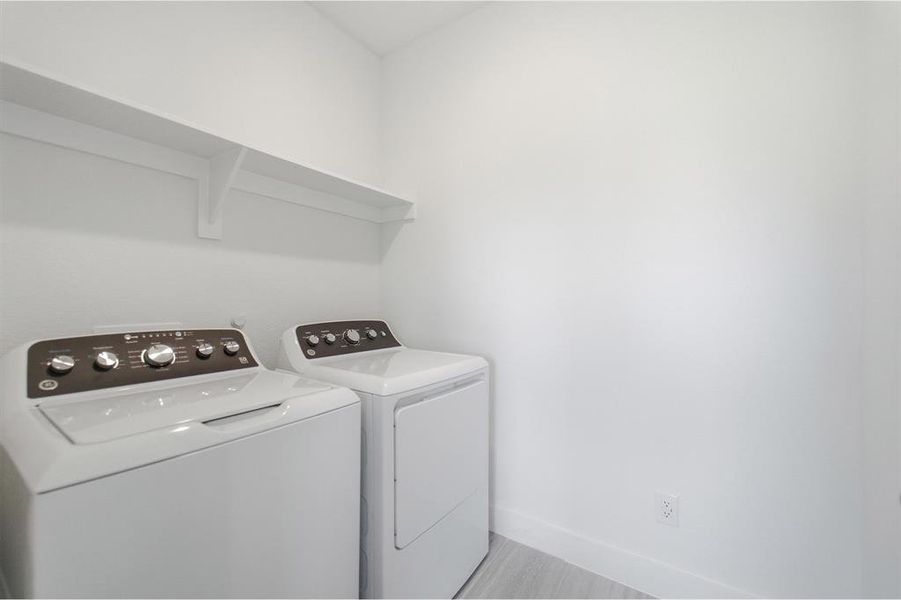 Dedicated laundry area featuring white walls, a white wall-mounted shelf, and light wood-finish flooring Dedicated laundry area featuring white walls, a white wall-mounted shelf, and light wood-finish flooring