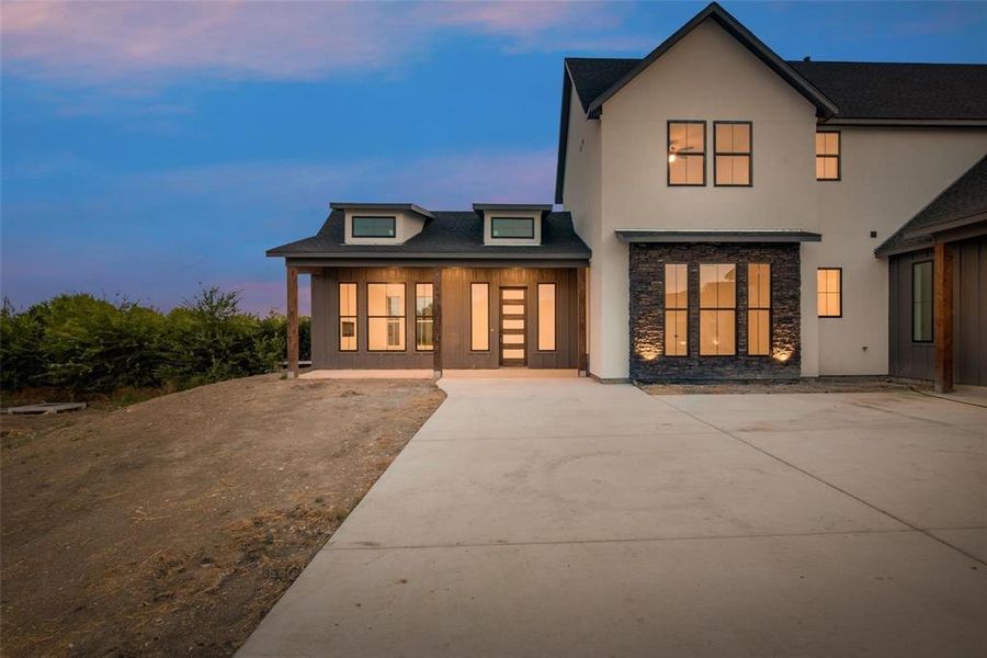 Back of house at dusk with stone siding, a porch, driveway, stucco siding, and a shingled roof