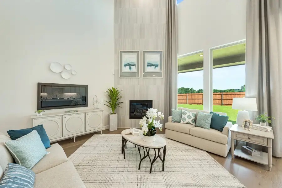Living room with two-story ceiling, fireplace, large windows, and neutral furniture with teal accent pillows