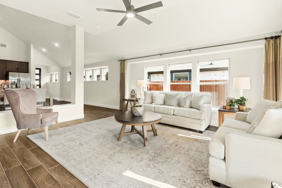 Open living room with white sofas, wood-look tile floors, ceiling fan, and large windows overlooking a backyard.