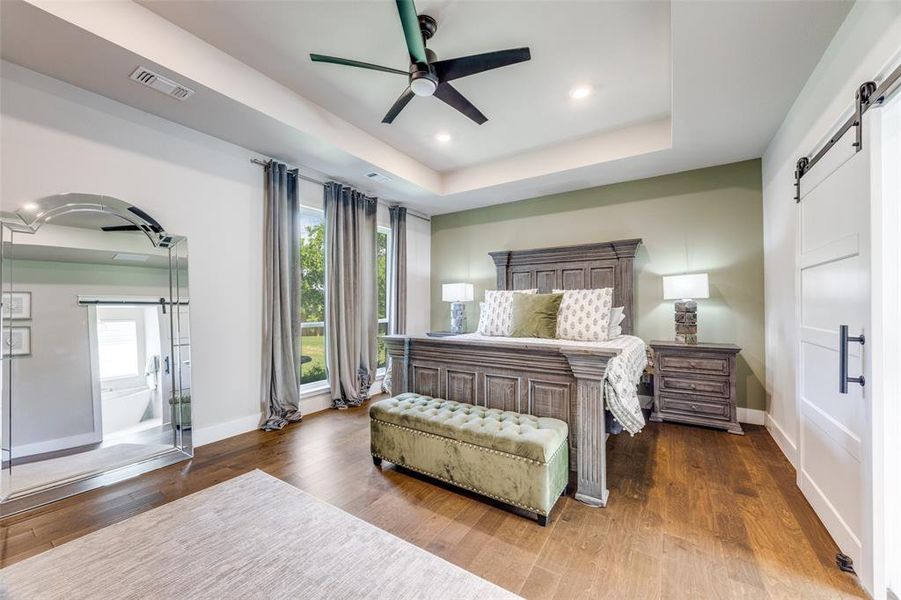 Bedroom with a barn door, a raised ceiling, multiple windows, dark wood-style flooring, and recessed lighting